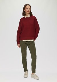 Burgunderfarbener Strickpullover mit gerippter Textur und umgeschlagenen Ärmeln, über einem beigen Hemd mit Kragen getragen; kombiniert mit olivgrünen, schmal geschnittenen Hosen und beigen Sneakers.