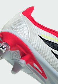 Valkoinen ja punainen jalkapallokengän nappulakengän pintamateriaali on teksturoitu, siinä on musta korostuslogo, näkyvät nastat pohjassa ja virtaviivainen muotoilu.