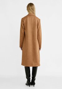 Mujer vistiendo un abrigo largo color camel y botas negras de tacón hasta la rodilla, de pie de espaldas contra un fondo blanco plano.