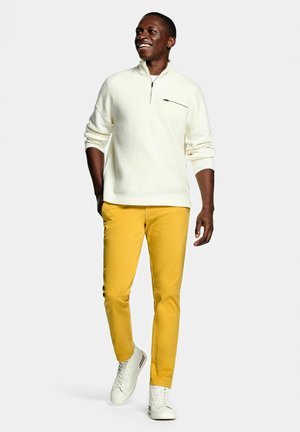 Lachende man wandelt, gekleed in een wit trui met kwart-ritssluiting, mosterdgele broek en witte sneakers tegen een effen achtergrond.