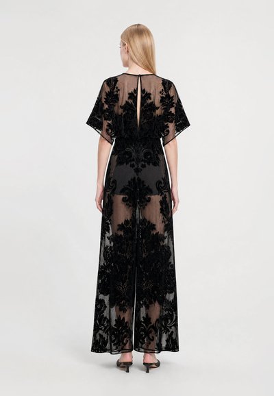Woman with long blonde hair wears a sheer black floral lace dress with wide sleeves and a slit at the back, paired with black heeled sandals.