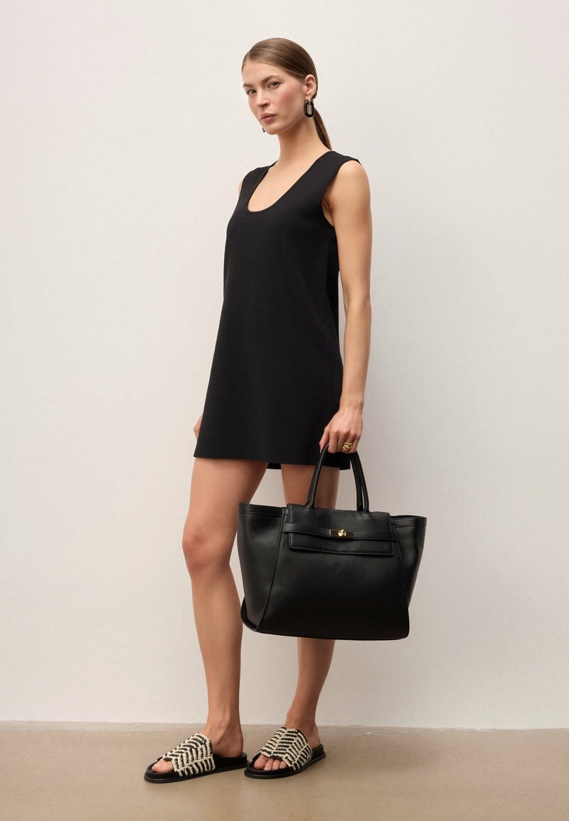 Femme en mini-robe noire sans manches, portant un grand sac à main noir, chaussée de sandales slip-on tissées noir et blanc, debout contre un mur uni.