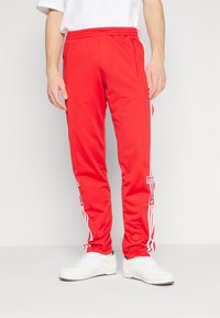 Muž v červených sportovních kalhotách Adidas s bílými pruhy a logy, v kombinaci s bílými teniskami, stojící na jednobarevném pozadí.