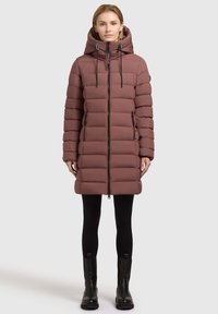 Pufīgs, vidēja garuma mētelis blāvi rozā krāsā ar rāvējslēdzēju priekšā, regulējamu kapuci un melniem akcentiem. Strukturēts, kvilts dizains; satur sānu kabatas.