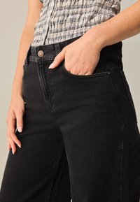 Sorte denimjeans har høj talje, vide ben og diskrete syninger. Modellens hånd hviler i sidelommen, hvor en kobberfarvet knapdetalje er synlig.