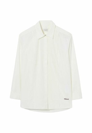 Femme portant une veste en cuir marron, une chemise blanche, une jupe plissée bleu marine, des chaussettes beiges et des baskets bordeaux, debout devant un fond uni.