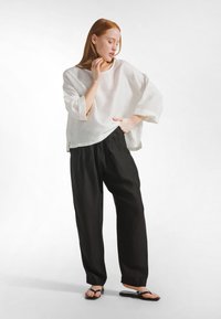 Kvinde med rødt hår iført en løs hvid bluse, sorte bukser med vide ben og sorte sandaler, stående med den ene hånd på hoften og øjnene lukkede.