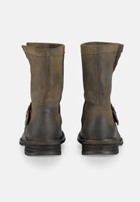 Botas de cuero marrón hasta el tobillo con una textura desgastada, un puño ligeramente doblado hacia abajo y hebillas laterales, con una suela de goma negra.