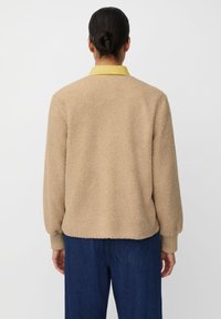 Beżowa kurtka z teksturowanej pluszowej tkaniny, posiadająca żółty kołnierz i ściągacze na mankietach, noszona na niebieskich dżinsach.