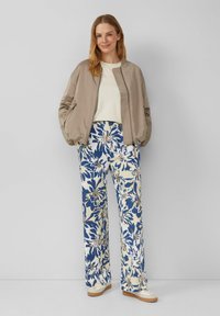 Beige bomberjack, witte top, bloemenbroek in blauw en crème met wijde benen en een gestructureerd patroon, gecombineerd met witte sneakers.