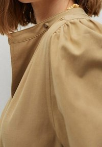 Gros plan d'une personne portant un vêtement beige avec un col montant, un détail de boutons et une épaule légèrement bouffante.