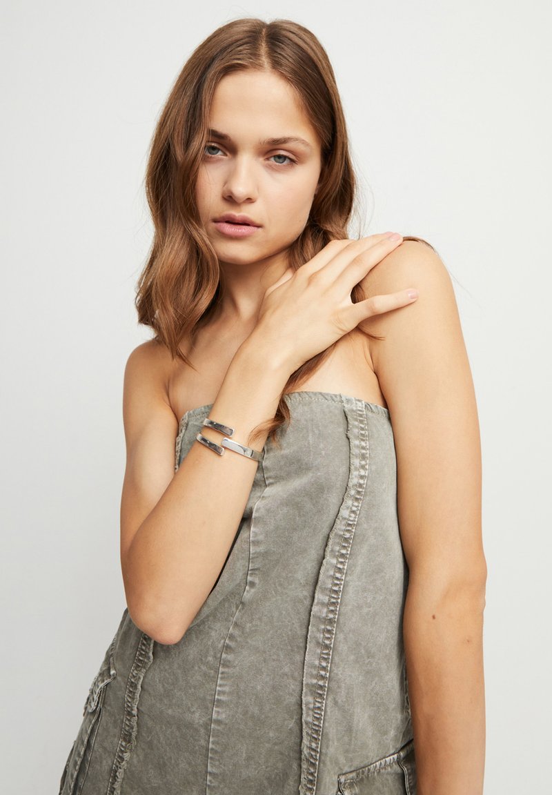 Robe sans bretelles couleur khaki avec une finition texturée et des poches latérales. Bracelet en métal argenté avec un design géométrique au poignet.