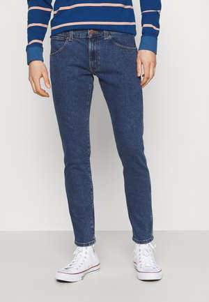 Blaue Denim-Jeans mit Slim-Fit, fünf Taschen-Design und leichter Waschtextur. Kombiniert mit weißen High-Top-Sneakers.