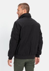 Black bomber jacket with a high collar, elastic cuffs, and waistband. Features a patch pocket on the left sleeve. Smooth, matte finish.