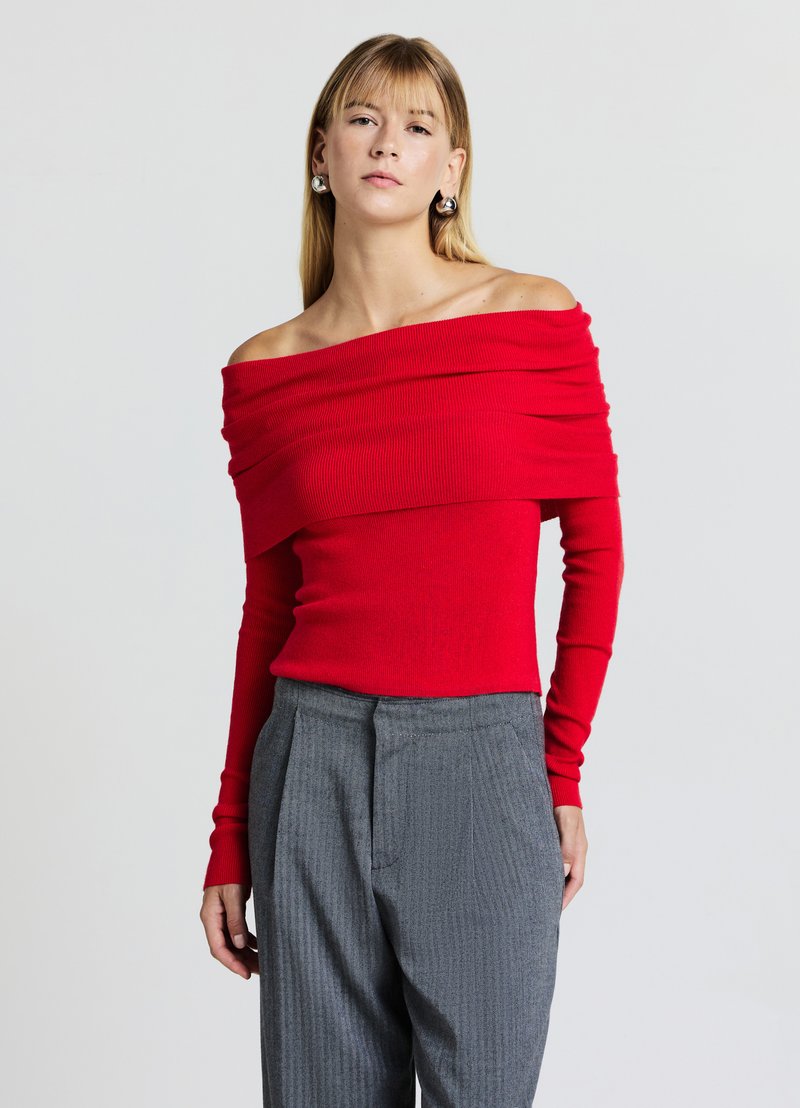 Roter, schulterfreier, gerippter Pullover mit langen Ärmeln, strukturierter Stoff, getragen mit grauen, nadelstreifenbesetzten Hosen. Elegantes Drapierungsdetail am Ausschnitt.