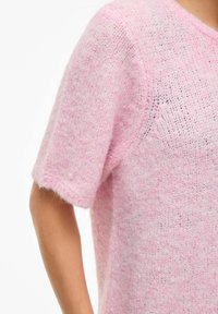 Rozā adīts džemperis ar īsām piedurknēm, kam raksturīga vaļēja adījuma struktūra un mīksta tekstūra, ar smalkām krāsas niansēm un brīvu piegriezumu.