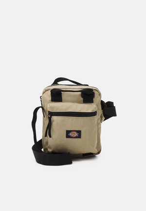 Kleine beige canvas schoudertas met voorvak met rits, zwarte verstelbare riem, bovenhandvat en Dickies-logo op het voorvak.