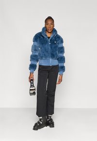 Casaco cropped de pelúcia azul com mangas largas, fecho de zip, bainha canelada, combinado com calças pretas e botas de ski, segurando óculos de ski.