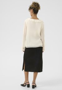 Lys beige bluse med flæsekrave og lange ærmer; parret med en sort knælang nederdel med en sideslids og sandaler med blokhæl.
