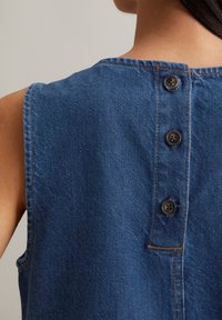 Denim mouwloze top met een blauwe stof, ronde halslijn en vier donkere knopen langs de achterkant, met zichtbare stikdetails.