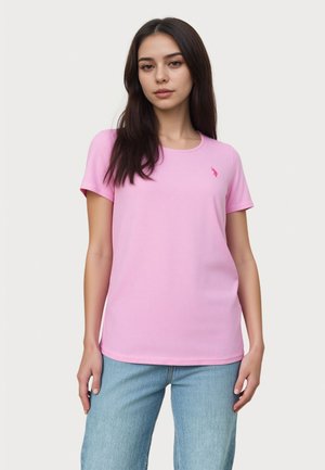 Jeune femme portant un t-shirt rose uni à manches courtes avec un petit logo sur la poitrine gauche, associé à un jean bleu clair, debout.