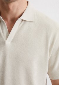 Lichtbeige polo shirt met een gebreide, gestructureerde stof, V-halsontwerp en een zachte kraag, wat een casual, minimalistische stijl benadrukt.