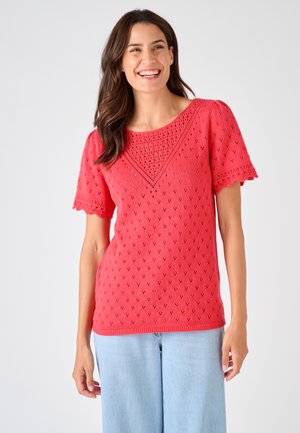 Mujer sonriendo que lleva un top de punto rojo de manga corta con detalles de encaje y vaqueros azul claro sobre un fondo claro y liso.