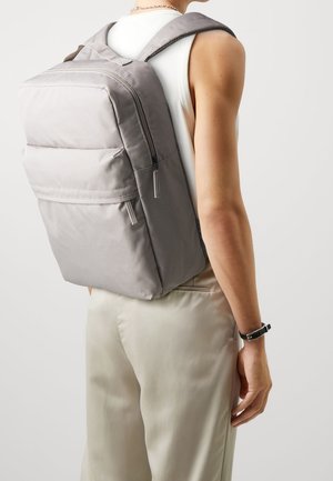 Mochila de tela gris con una forma estructurada, que cuenta con dos bolsillos frontales con cremallera y correas de hombro acolchadas para mayor comodidad. Diseño minimalista.