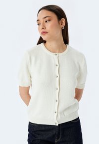 Cardigan bianco a maniche corte con scollatura rotonda, dotato di chiusura a bottoni e orlo a costine. Realizzato in morbido tessuto lavorato a maglia con una consistenza liscia.