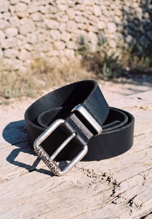Un cinturón de cuero negro con una hebilla plateada reposa sobre madera desgastada al aire libre, junto a un muro de piedra.