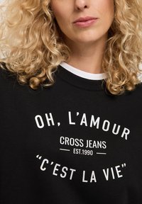 Czarny sweter z okrągłym dekoltem, z białym nadrukiem: "OH, L'AMOUR CROSS JEANS EST.1990 'C'EST LA VIE'." Tekst ma zakrzywiony układ.