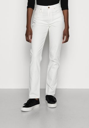 Person trägt weiße Straight-Leg-Jeans, schwarzen Langarm-Top und schwarze Sneakers mit weißen Sohlen, steht auf grauem Boden.
