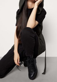 Schwarze Schnür-Kampfstiefel, schwarze eng anliegende Hosen und eine lange, dunkelgrüne Kapuzenweste mit glattem Textureffekt und verstellbaren Bändern.