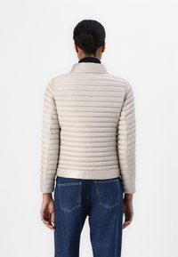 Beige dunjakke med horisontal søm, ståkrage og lange ermer, parret med blå denim jeans med baklommer.