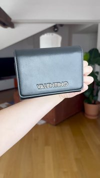 Cartera de piel negra con cierre magnético, que presenta un logotipo plateado "VALENTINO" en el frontal, forma rectangular y textura suave.