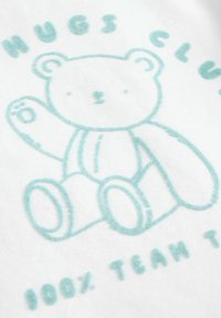 Fehér anyag, világoskék medve grafikával, kiemelkedő körvonalakkal. A szöveg: "ÖLELÉS KLUB" és "100% CSAPATTAG TEA." Puha tapintású.