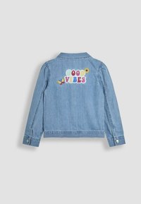 Chaqueta de denim azul claro con cuello, mangas largas y bordado "BUENAS VIBRAS" con acentos coloridos como una mariposa y una flor.
