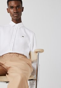 Chemise blanche à manches longues avec un logo brodé vert, associée à un pantalon beige. La chemise possède un col et une fermeture à boutons.