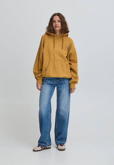 Dzeltena oversize kapuce ar kangaroo kabatu un saites, kopā ar gaiši ziliem plašajiem džinsiem un krēms krāsas kedām.