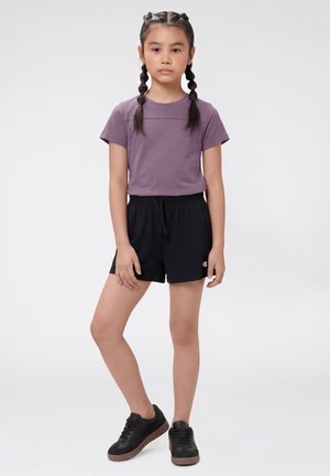 Jong meisje met gevlochten haar, een paarse T-shirt met korte mouwen, zwarte short met trekkoord en zwarte sneakers, staand met één been gekruist.