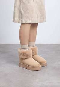 Botas de tobillo beige con un puño de piel de peluche, hechas de ante. Tienen una suela plana y texturizada y están combinadas con calcetines de punto crema.