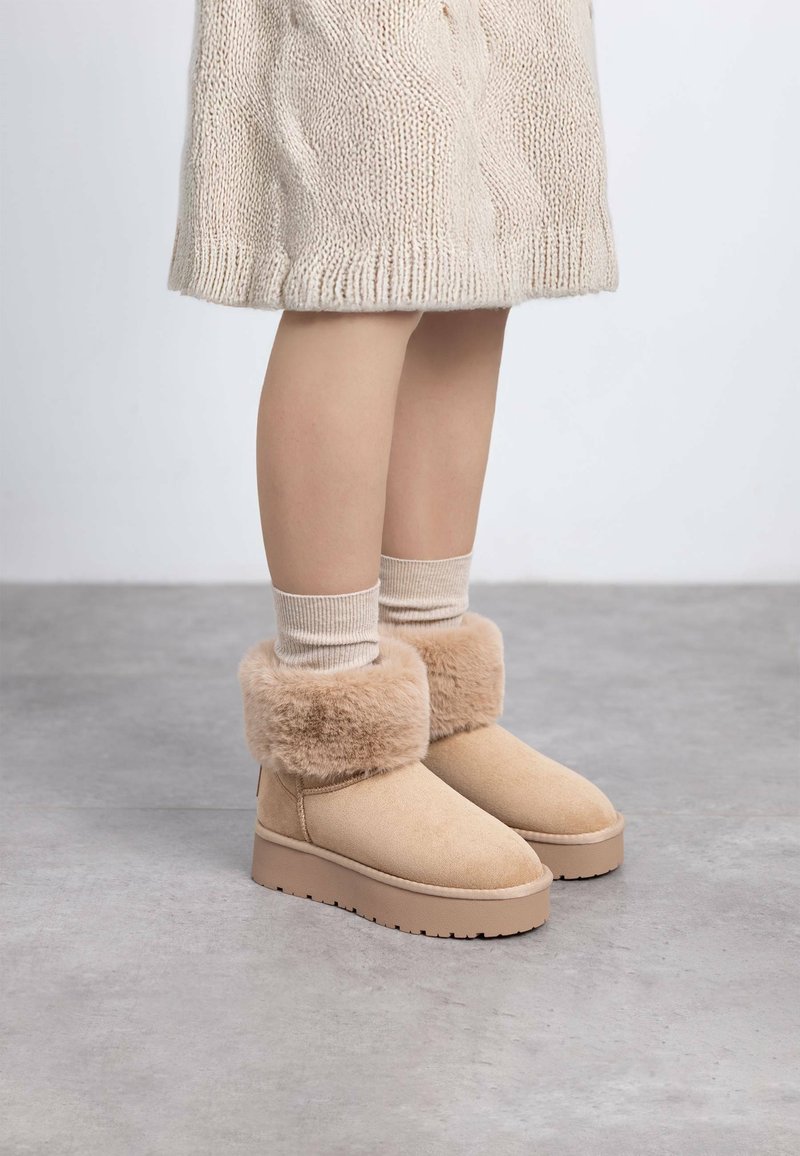 Botas de tobillo beige con un puño de piel de peluche, hechas de ante. Tienen una suela plana y texturizada y están combinadas con calcetines de punto crema.
