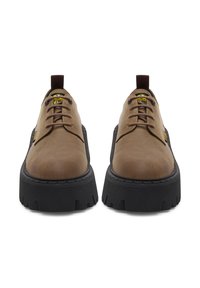 Zapatos de cuero marrón con punta redonda, suela de goma negra y patrones de dibujo prominentes. Presentan un diseño con cordones y una etiqueta de marca amarilla.