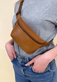 Braune Leder-Hüfttasche mit einer geschwungenen Form, vorderer Reißverschlusstasche und verstellbarem Riemen, getragen über einem grauen Shirt und blauen Jeans.