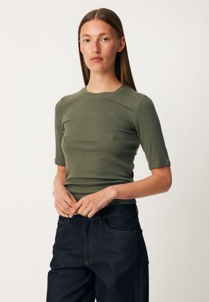 Femme aux cheveux bruns lisses portant un haut vert à manches courtes et un jean bleu foncé, debout devant un fond blanc uni.