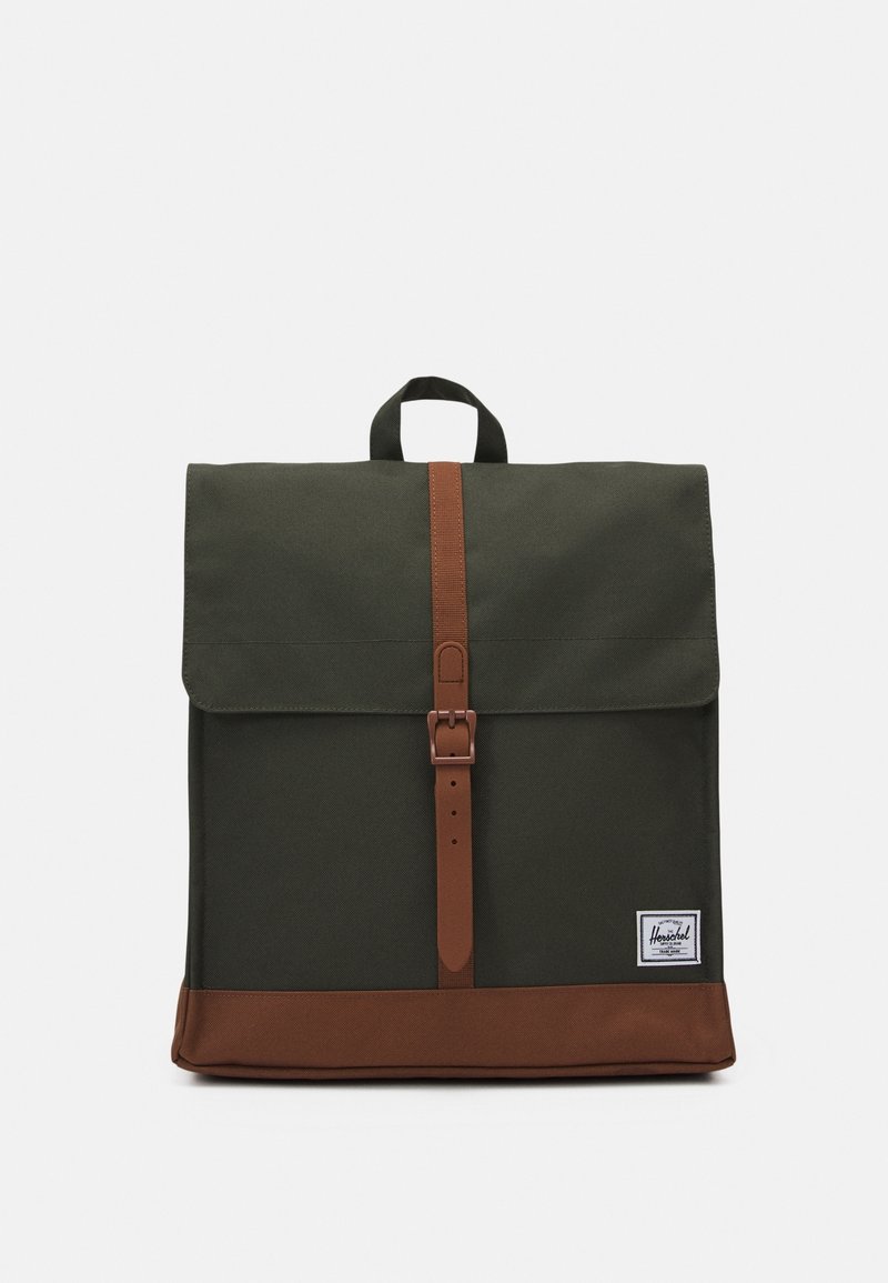 Zielony plecak z materiału z brązowymi skórzanymi akcentami, z zapięciem na guzik i uchwytem na górze. Posiada małą metkę marki na dole.