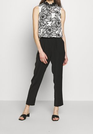 Kobieta w czarnych, wysokich, zwężanych spodniach, białym, bezrękawnym topie z czarnym kwiatowym wzorem oraz czarnych sandałkach na obcasie z odkrytymi palcami, stojąca.