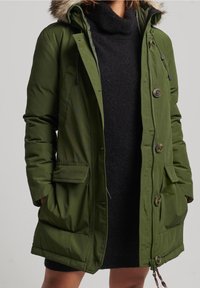 Vrouw die een open, olijfgroene gewatteerde parka met een capuchon met bontkraag draagt, over een zwart gebreide jurk, met haar handen in de zakken van de jas.