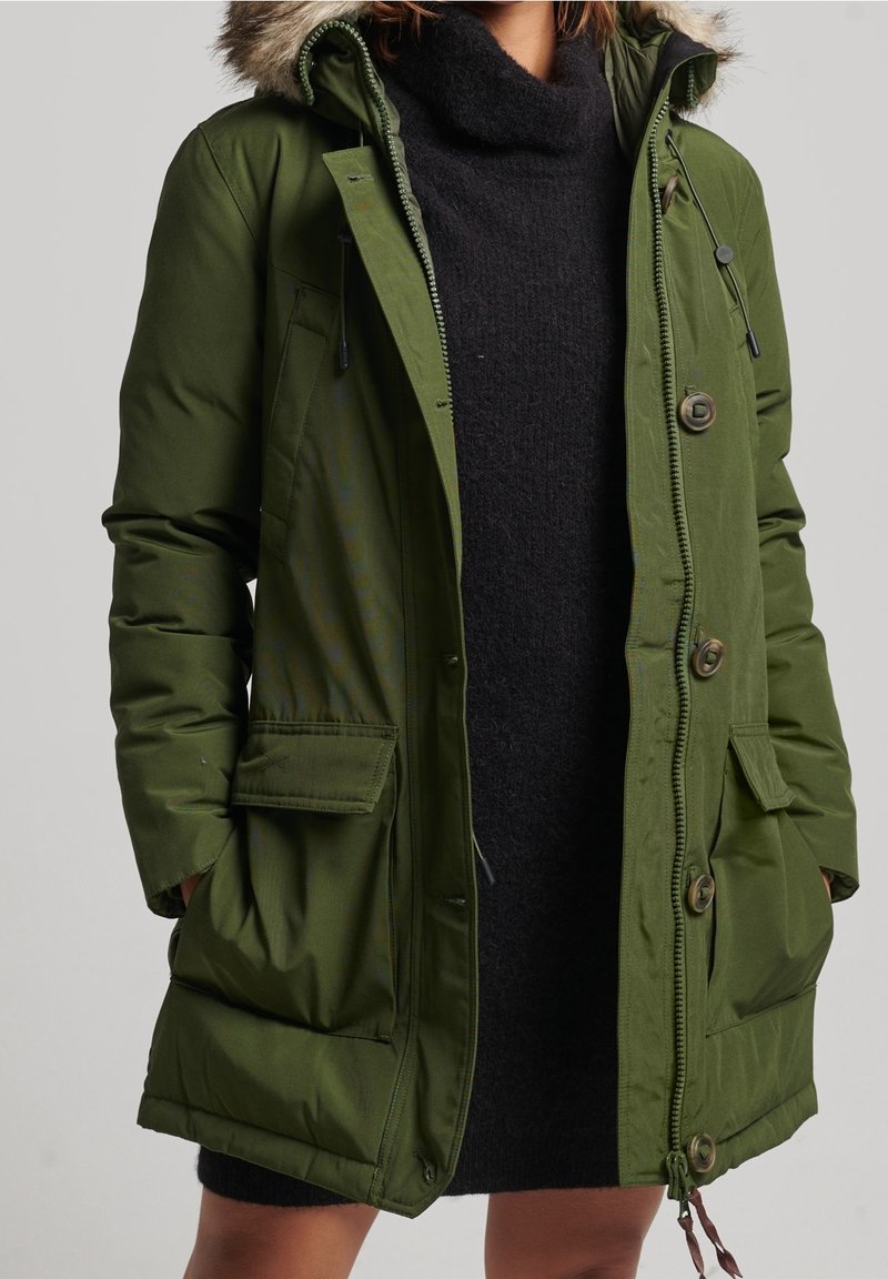 Vrouw die een open, olijfgroene gewatteerde parka met een capuchon met bontkraag draagt, over een zwart gebreide jurk, met haar handen in de zakken van de jas.