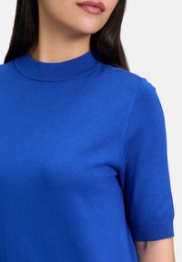 Top blu corto a maniche corte con collo alto, realizzato in tessuto morbido. Presenta colletto e orli delle maniche a costine. Colore solido, senza fantasie.